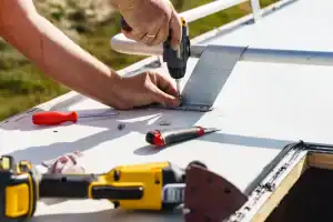 A person uses a cordless power drill to secure a screw into a white metal surface, with various tools like a screwdriver, wrench, and sanding tool lying nearby on the surface.