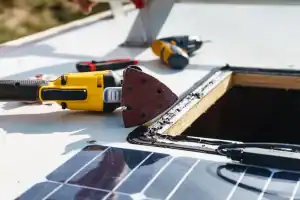 A close-up of tools, including a yellow electric sander and a screwdriver, on a white surface near a rectangular cut-out and adhesive sealant, with solar panels partially visible.
