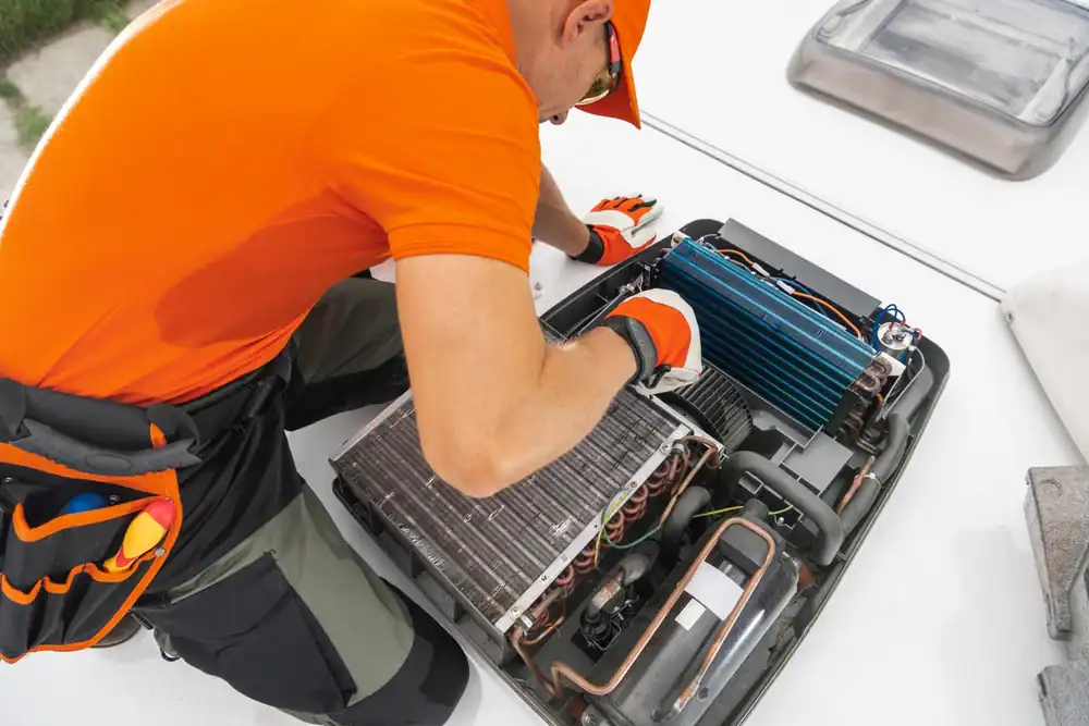 A technician in an orange shirt and cap repairs the exposed interior of an air conditioning unit, using tools from a belt while working outdoors.